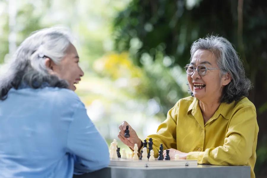 Two senior women enjoy a chess game outdoors surrounded by nature.